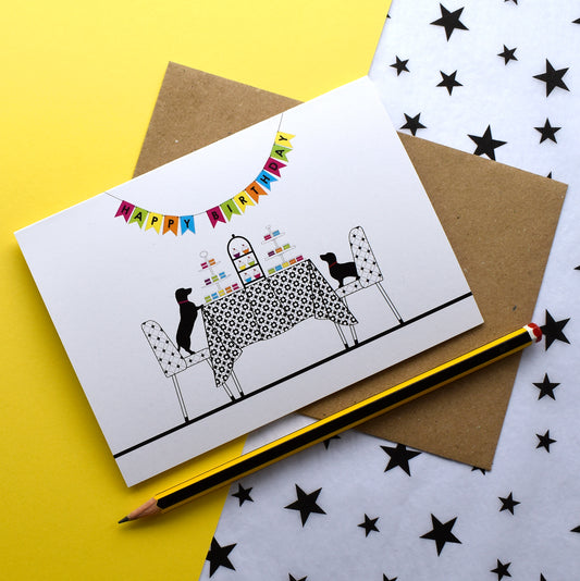 A collection of colourful greeting cards featuring dachshunds, Mali & Penny and their adventures.  This English birthday card shows them at a birthday party, sitting at a table full of cake!